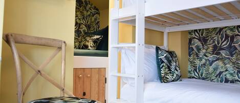 Ein Etagenbett mit weißer Bettwäsche, ein Holzhocker mit gemustertem Sitzkissen und eine Wand mit tropischem Blattmuster.