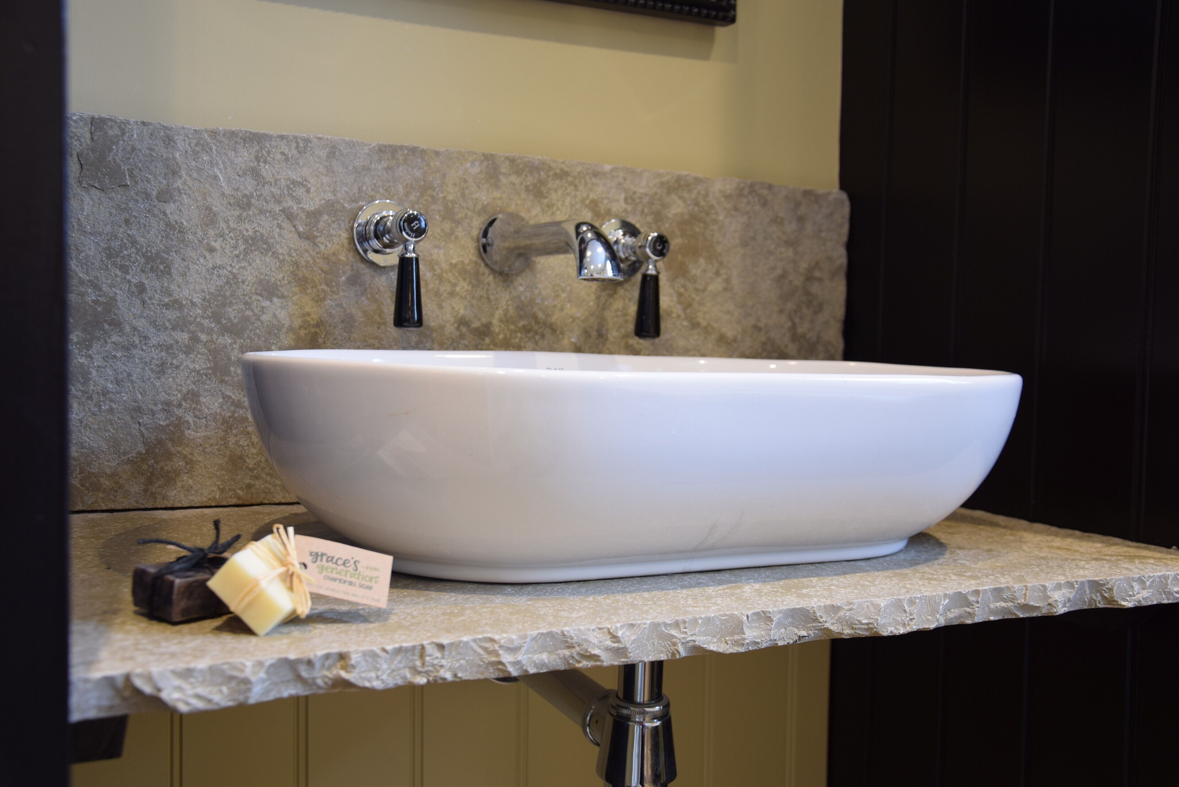 A bathroom with a large white vessel sink, two chrome faucets, and a stone-textured wall.