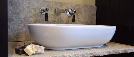 A bathroom with a large white vessel sink, two chrome faucets, and a stone-textured wall.