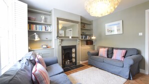 A living room with a fireplace, grey sofas, a bookshelf, and a chandelier. - 15 Victoria Street, Southwold (Reydon)