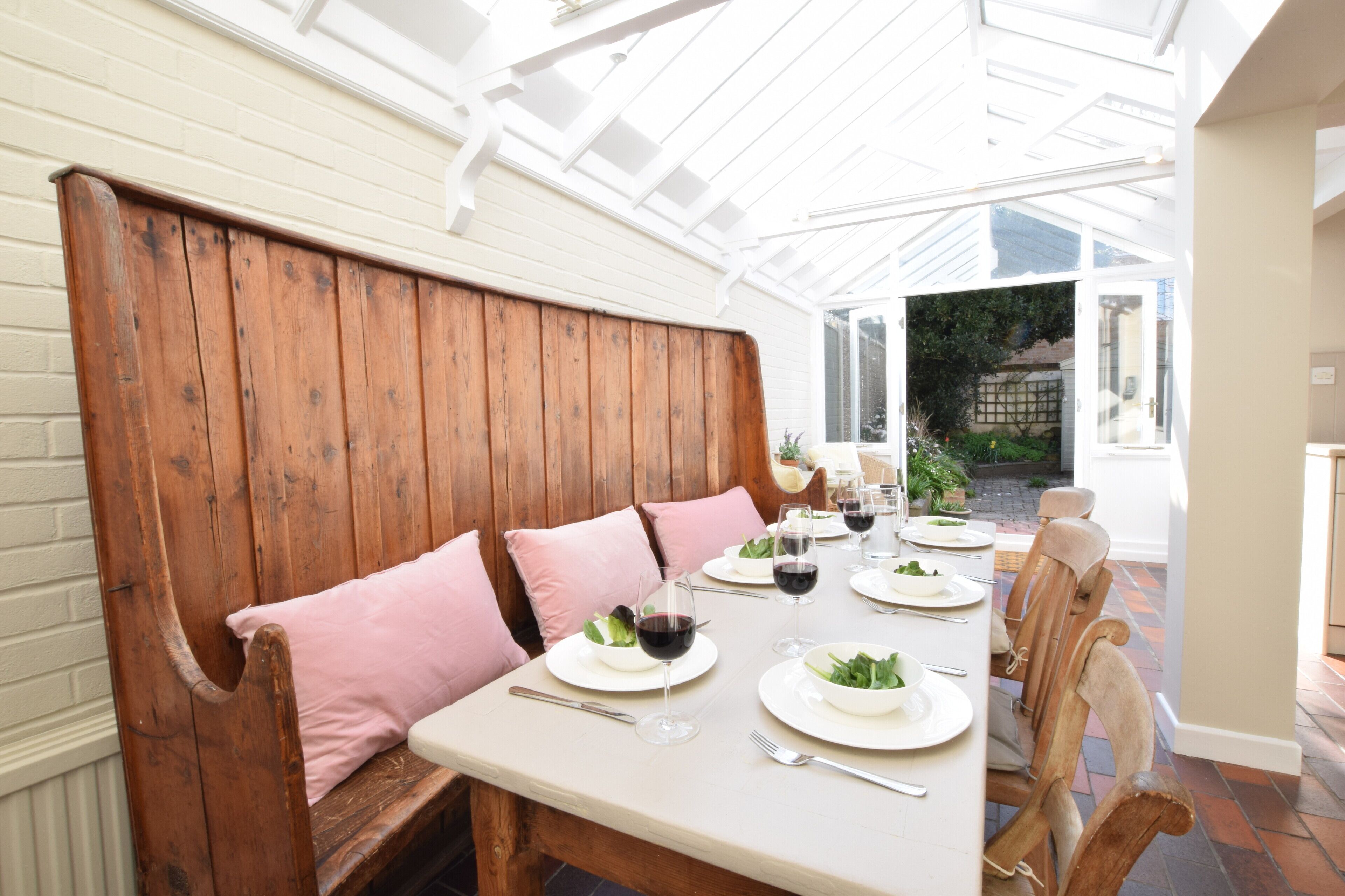 Un coin repas avec un banc en bois, une table blanche et des coussins roses.