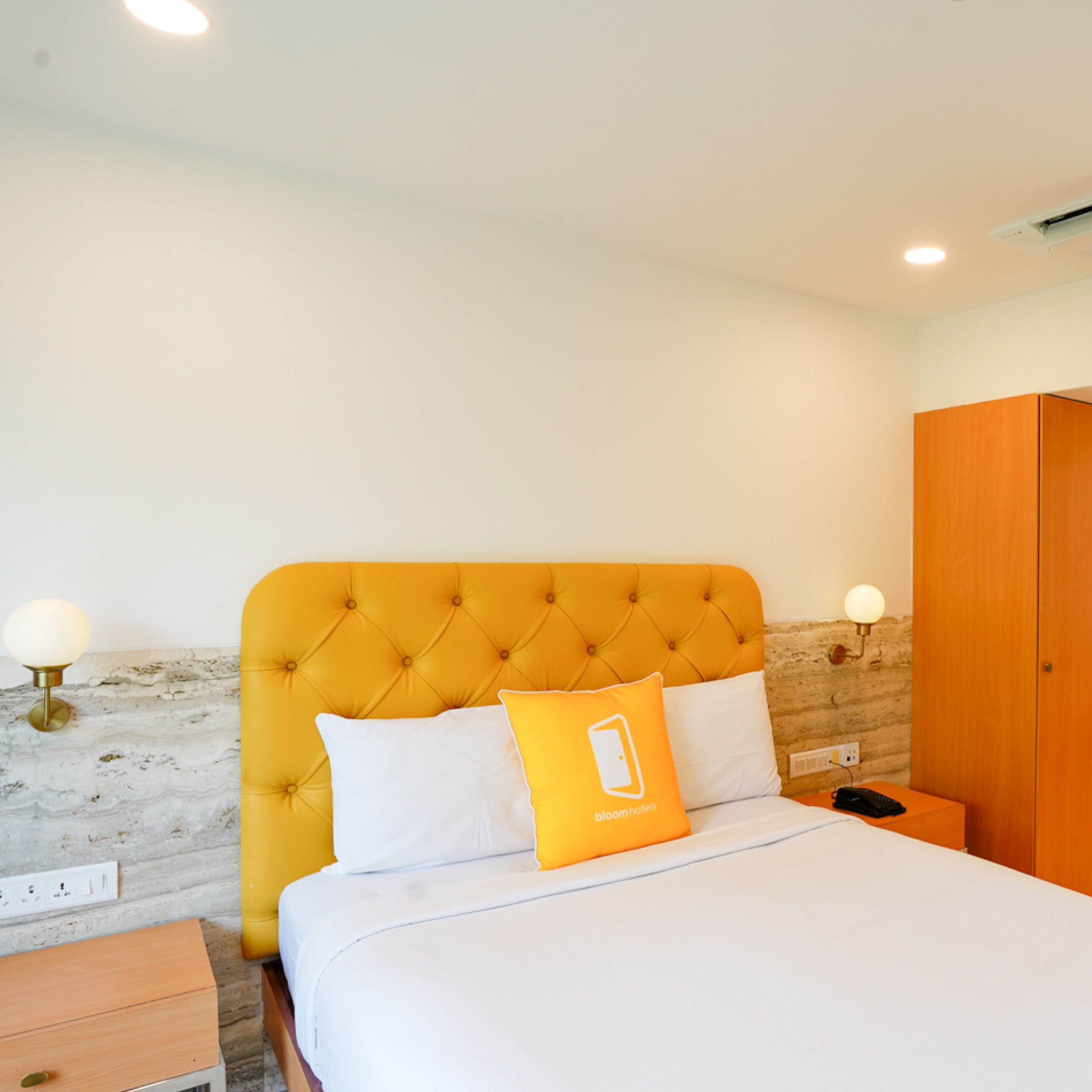 Une chambre d’hôtel avec une tête de lit capitonnée, un lit avec une literie blanche et orange, une table de chevet en bois et une armoire en bois.