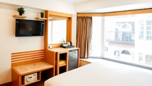 A hotel room with a bed, a desk with a chair, a television mounted on the wall, a window with curtains, and a small shelf with a plant. - Bloom Boutique - Bandra (Mumbai)