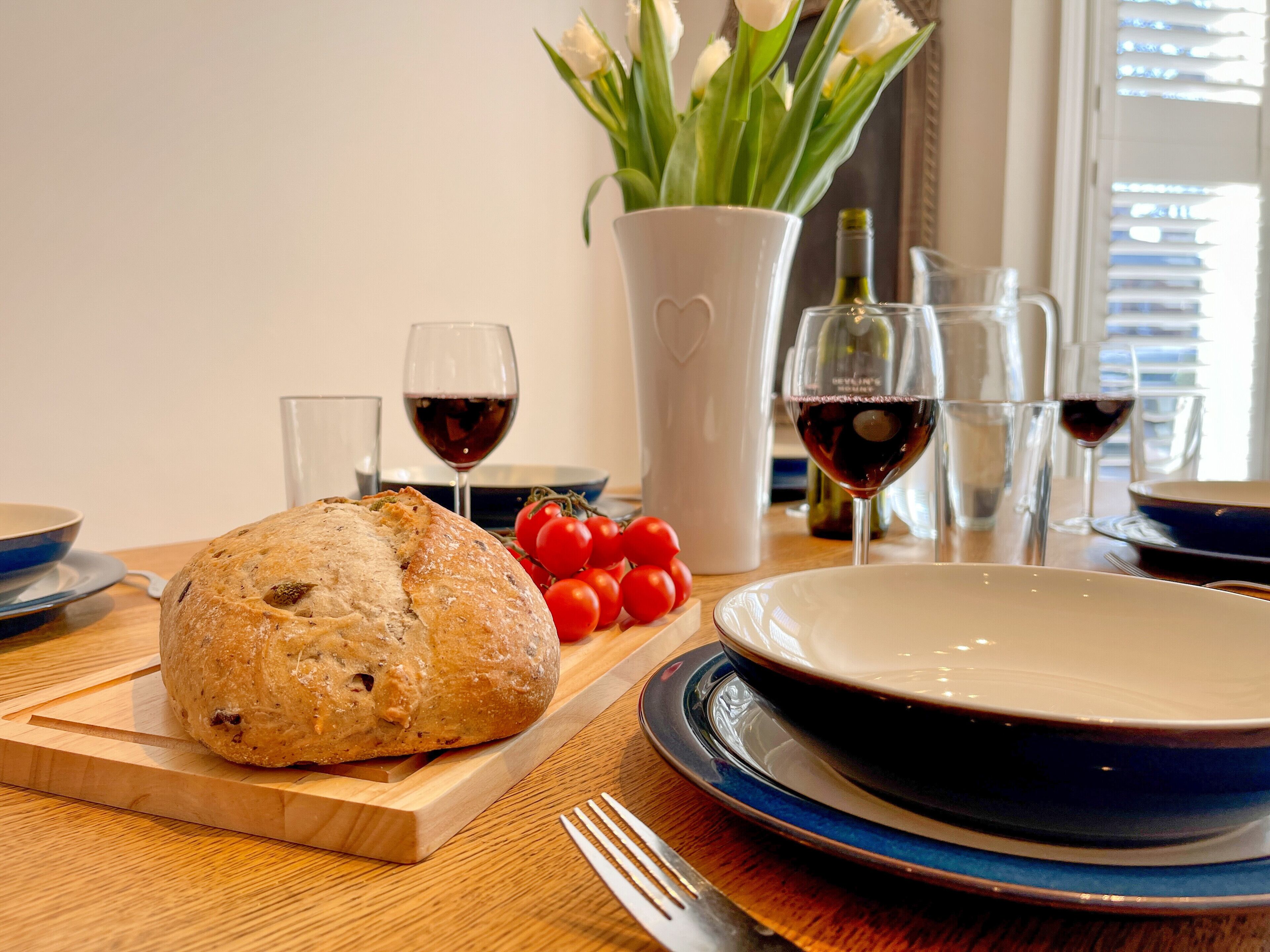 A dining table with a loaf of bread, a plate, a vase with flowers, and glasses of wine.