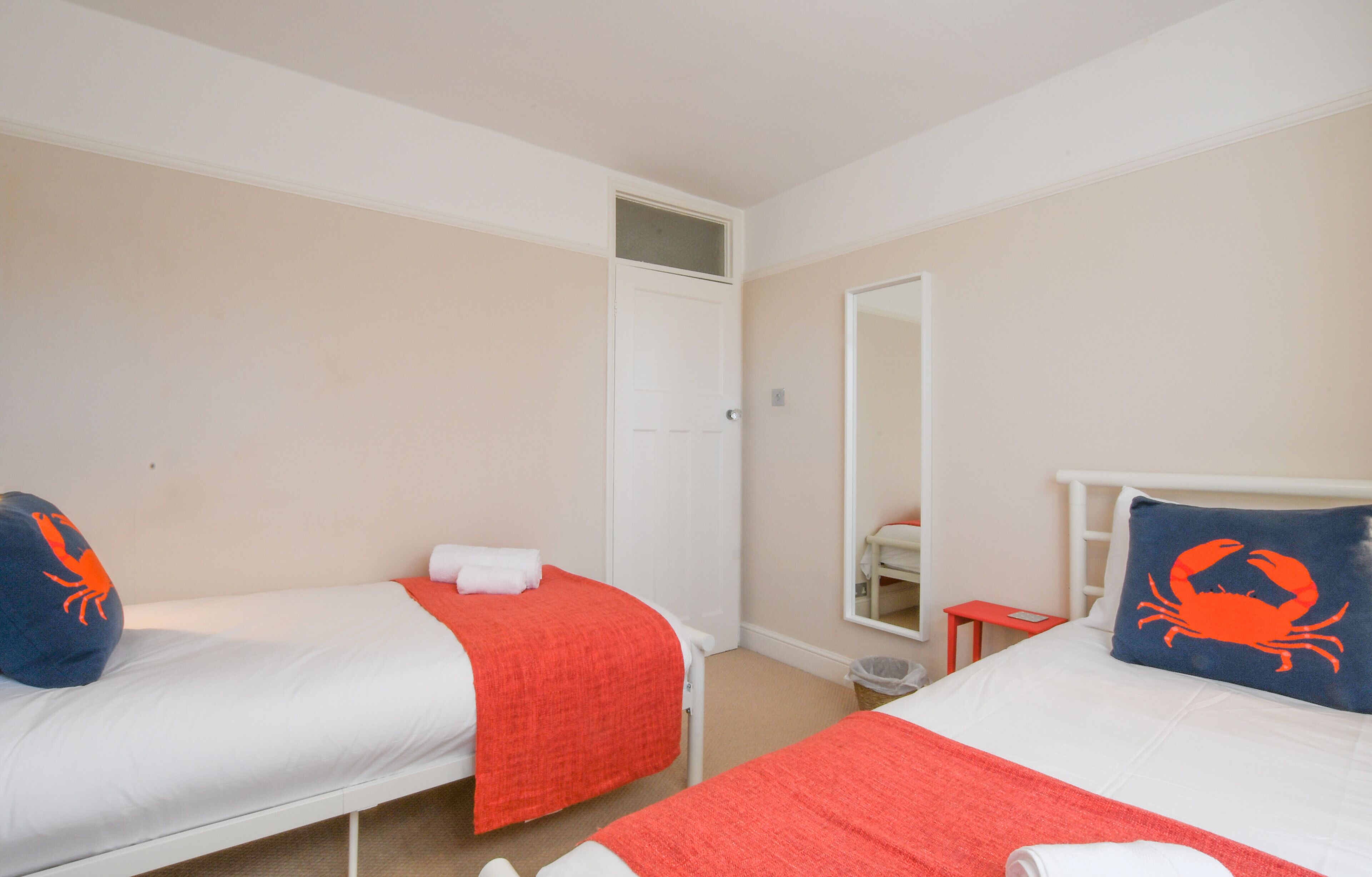 A hotel room with two single beds, each with a red blanket and a blue pillow with a crab design. The room has beige walls, a white door, and a small red table.