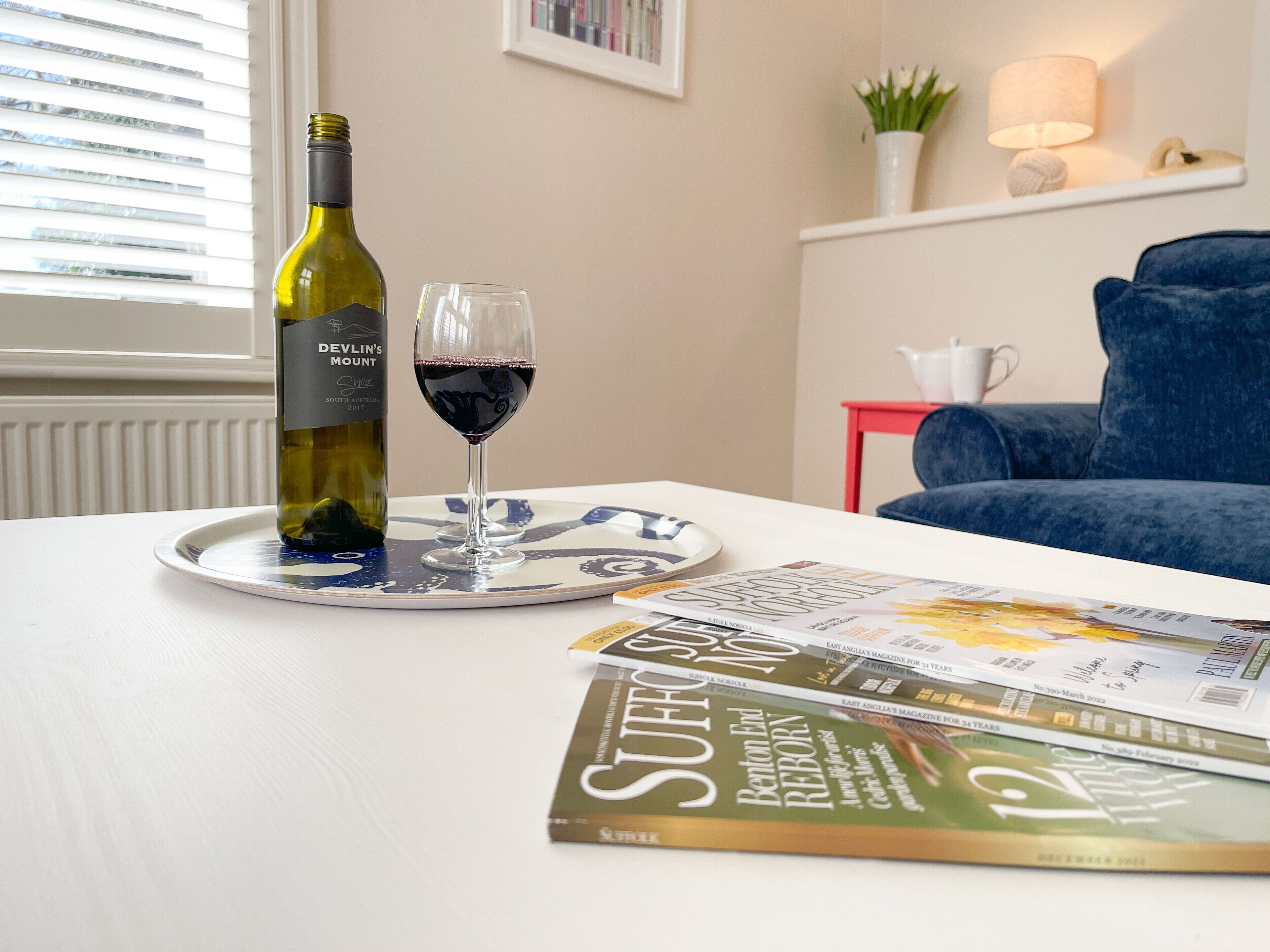 Uma garrafa de vinho e um copo em um suporte, uma revista e uma cadeira azul em um quarto com janela e luminária.