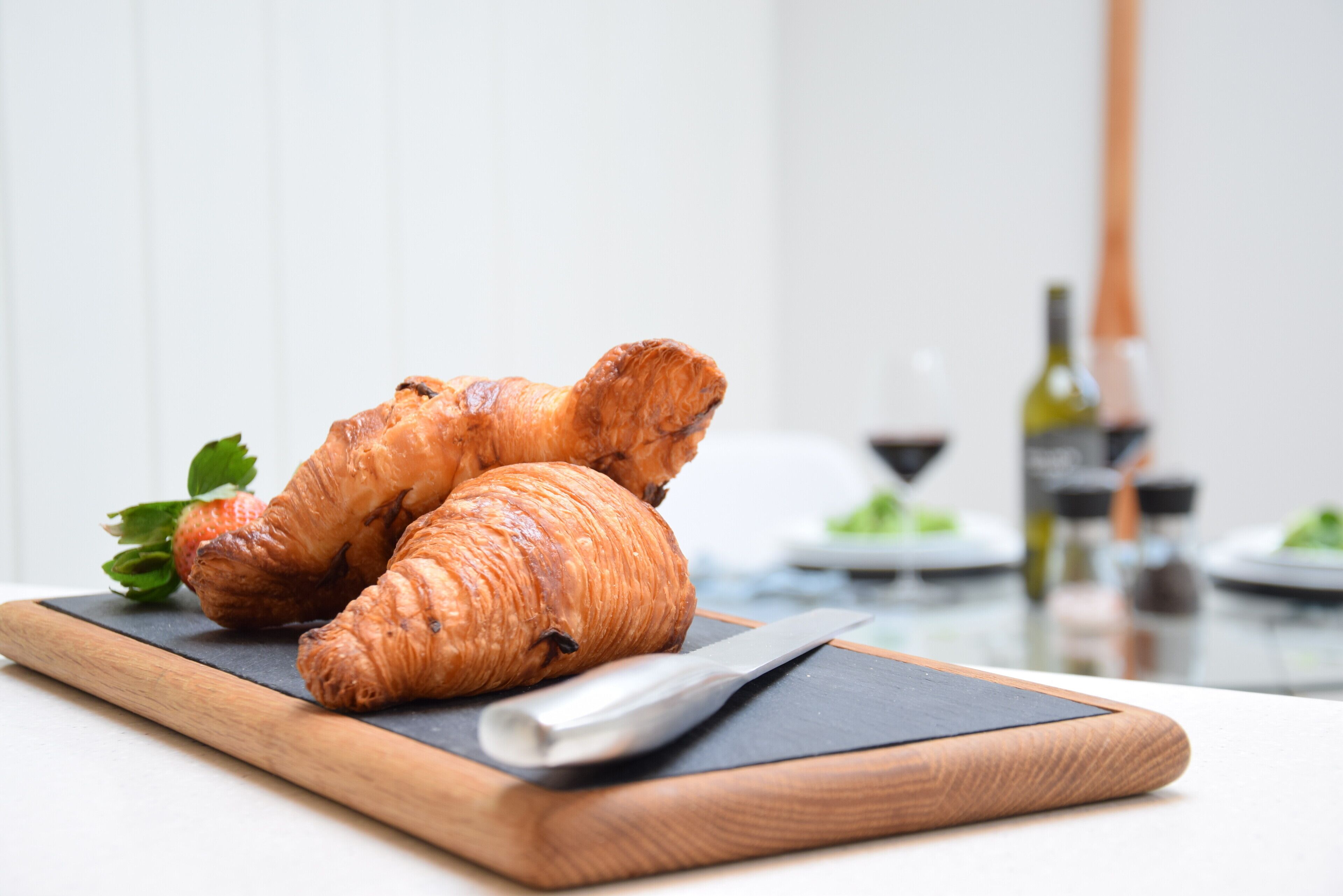 Des croissants sur une planche en bois, avec un couteau à côté, une bouteille de vin et un verre de vin rouge en arrière-plan.