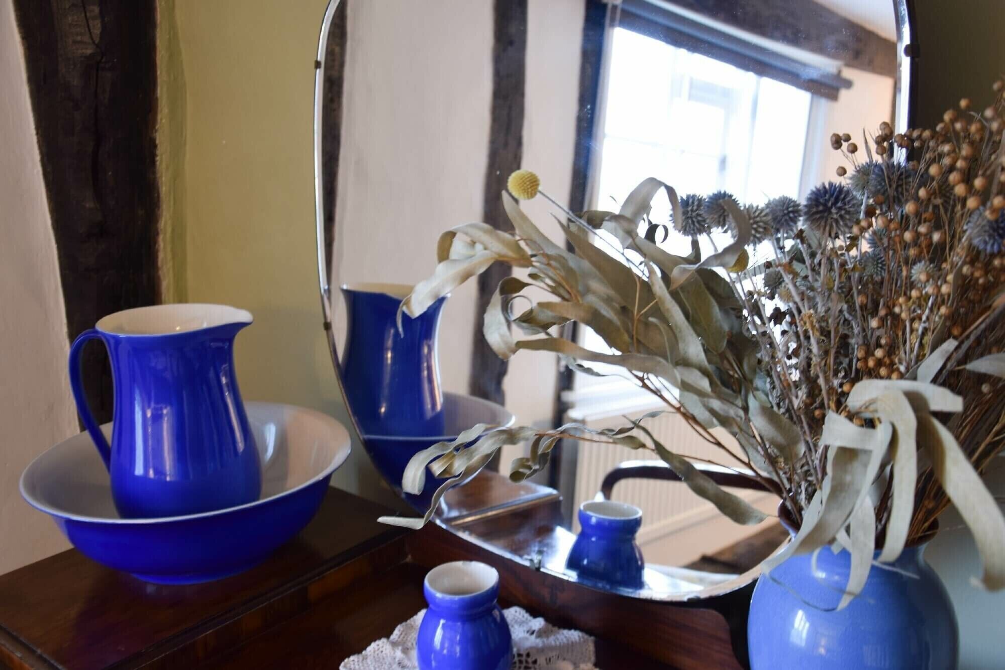 A mirror reflecting a blue ceramic pitcher, a blue bowl, and a vase with dried flowers.