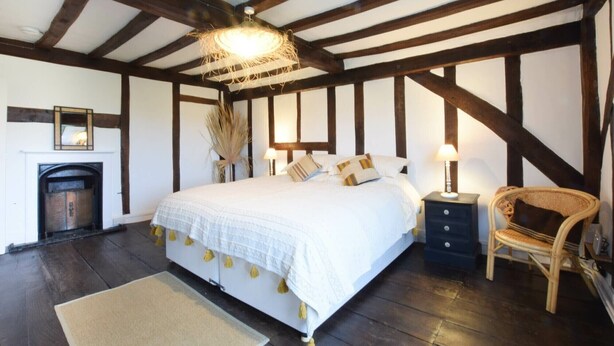 A bedroom with a large bed, a chair, a nightstand, a mirror, and a fireplace. - Church Farmhouse, Cookley (Halesworth)