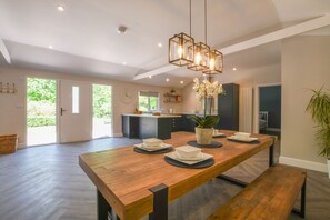 A spacious dining area with a long wooden table, a hanging light fixture, and a view of the kitchen and garden through the open door. - The Piglets (Woodbridge)