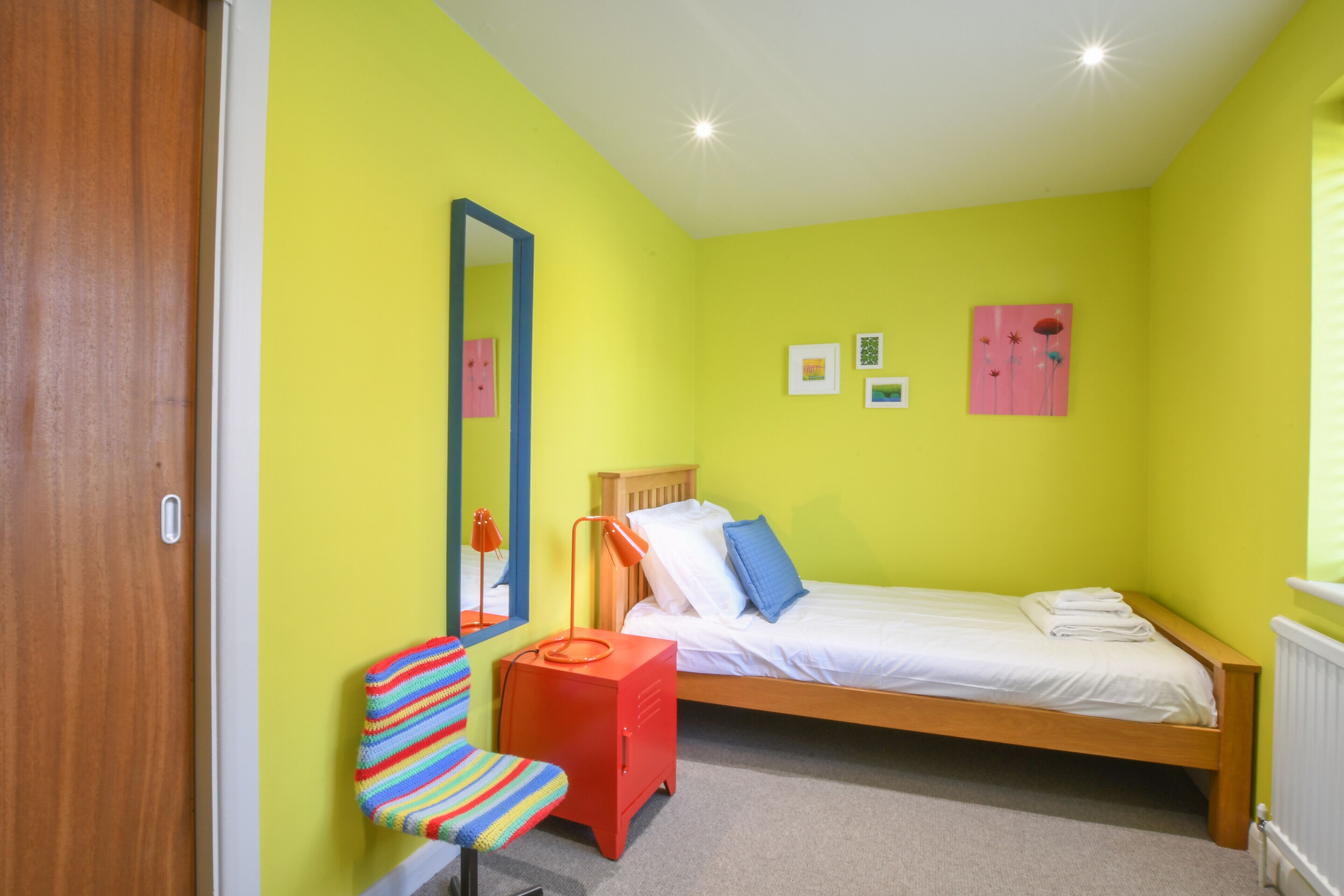 A bedroom with a single bed, a chair, a desk, and a mirror.