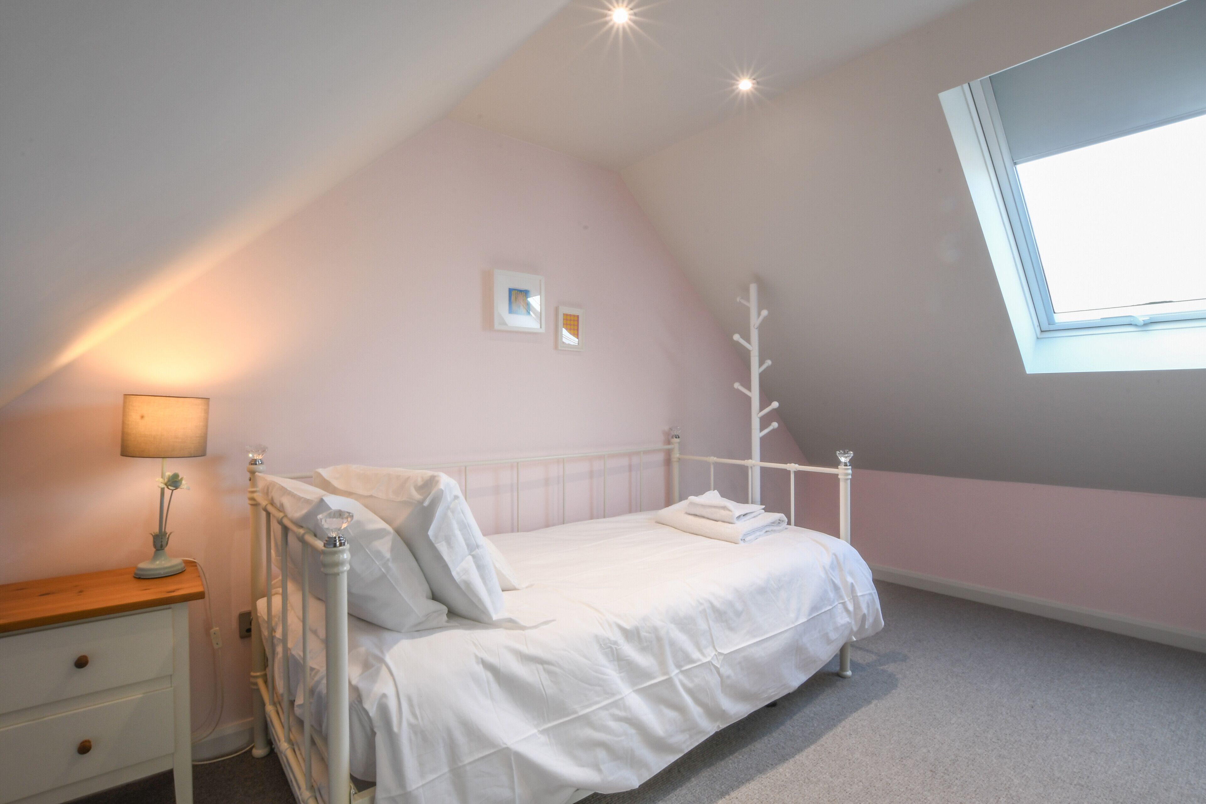 Une chambre avec un plafond en pente, un lit recouvert de draps blancs, une table de chevet avec une lampe et une fenêtre donnant sur le ciel.