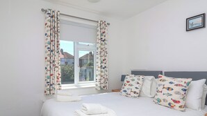 A neatly made bed with white linens and a fish-patterned pillow, a window with curtains, and a framed picture on the wall. - Time Out, Southwold (Reydon)