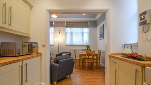 A compact kitchen with wooden cabinets, a microwave, and a dining area with a table and chairs. - 1 Eversley Court, Southwold (Reydon)