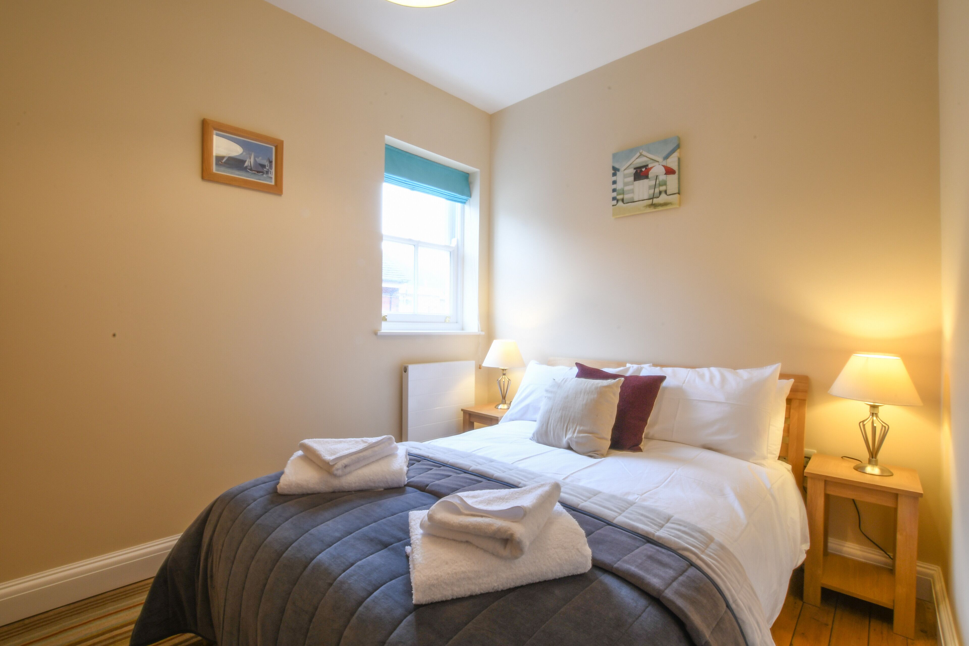 Ein ordentlich gemachtes Bett mit Kissen und Decke, zwei Nachttische mit Lampen, ein Fenster mit blauem Vorhang und zwei gerahmte Bilder an der Wand.