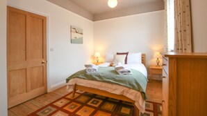 A bedroom with a wooden door, a bed with a green blanket, two bedside lamps, and a nightstand with a lamp. - 1 Eversley Court, Southwold (Reydon)
