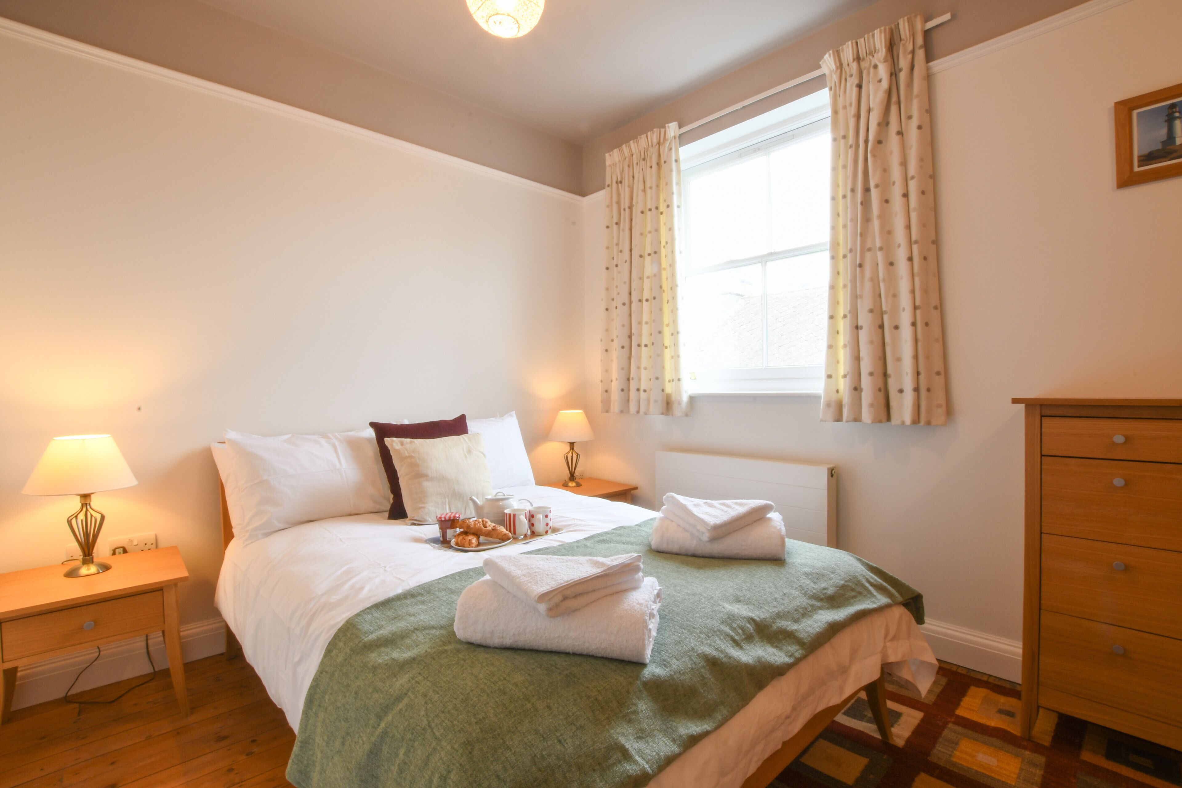 A neatly made bed with a green blanket, two bedside lamps, and a window with curtains.