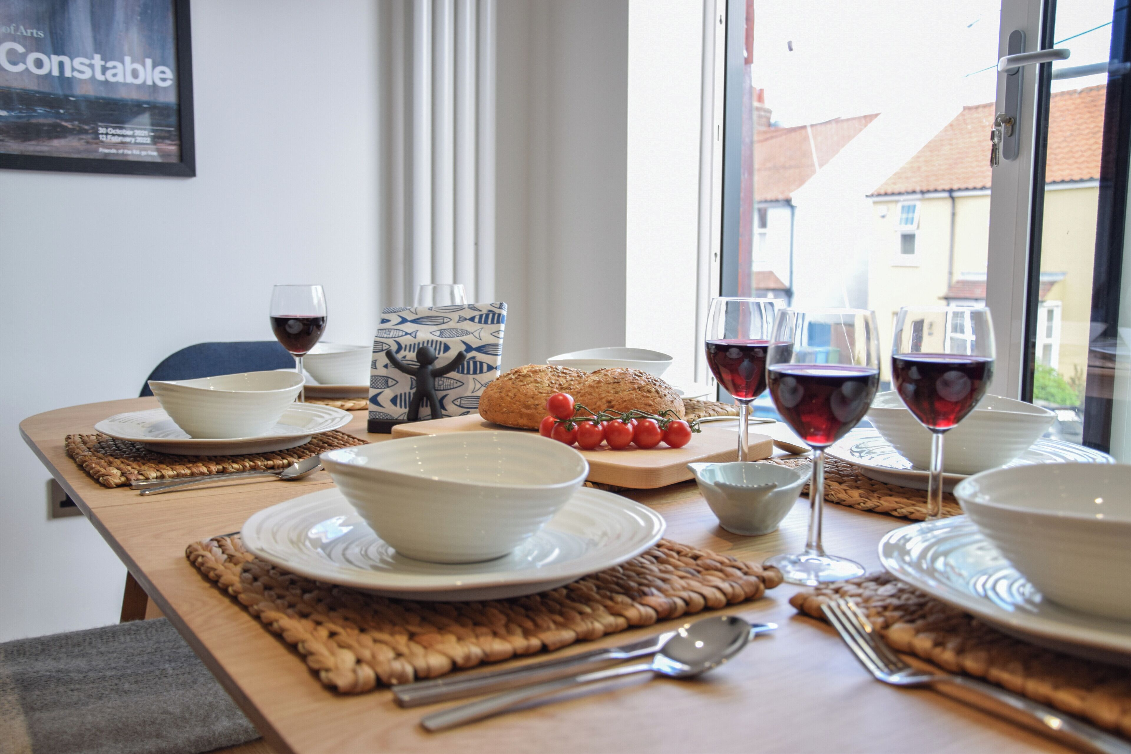 A dining table set with white bowls, plates, and cutlery, a loaf of bread, cherry tomatoes, and wine glasses filled with red wine.