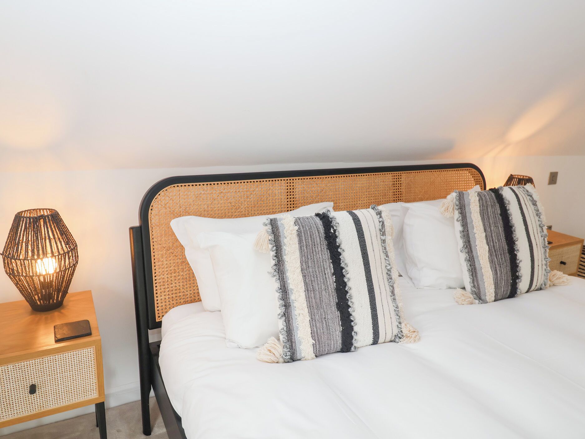Cama bem arrumada com almofadas listradas, cabeceira trançada e abajur sobre mesa de cabeceira de madeira.