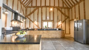 A kitchen with exposed wooden beams, a central island with a bowl of fruit, a stainless steel refrigerator, and a window with a view. - Halcyon Barn, Wissett (Halesworth)