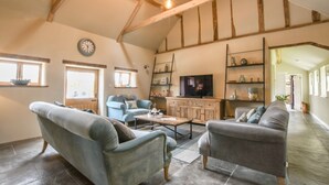 A spacious living room with high ceilings and exposed beams, featuring a large television, a fireplace, and a cozy seating area with two sofas and a coffee table. - Halcyon Barn, Wissett (Halesworth)