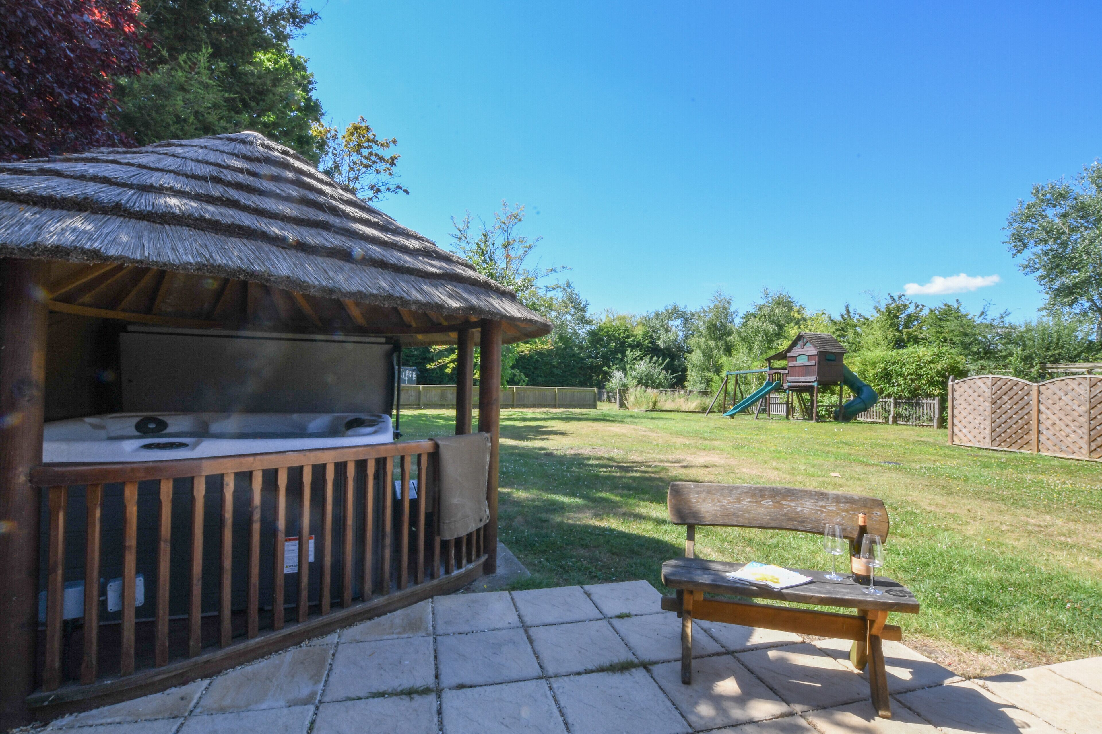 Un jacuzzi con una terraza de madera y un banco de madera en un patio trasero con una zona de juegos al fondo.