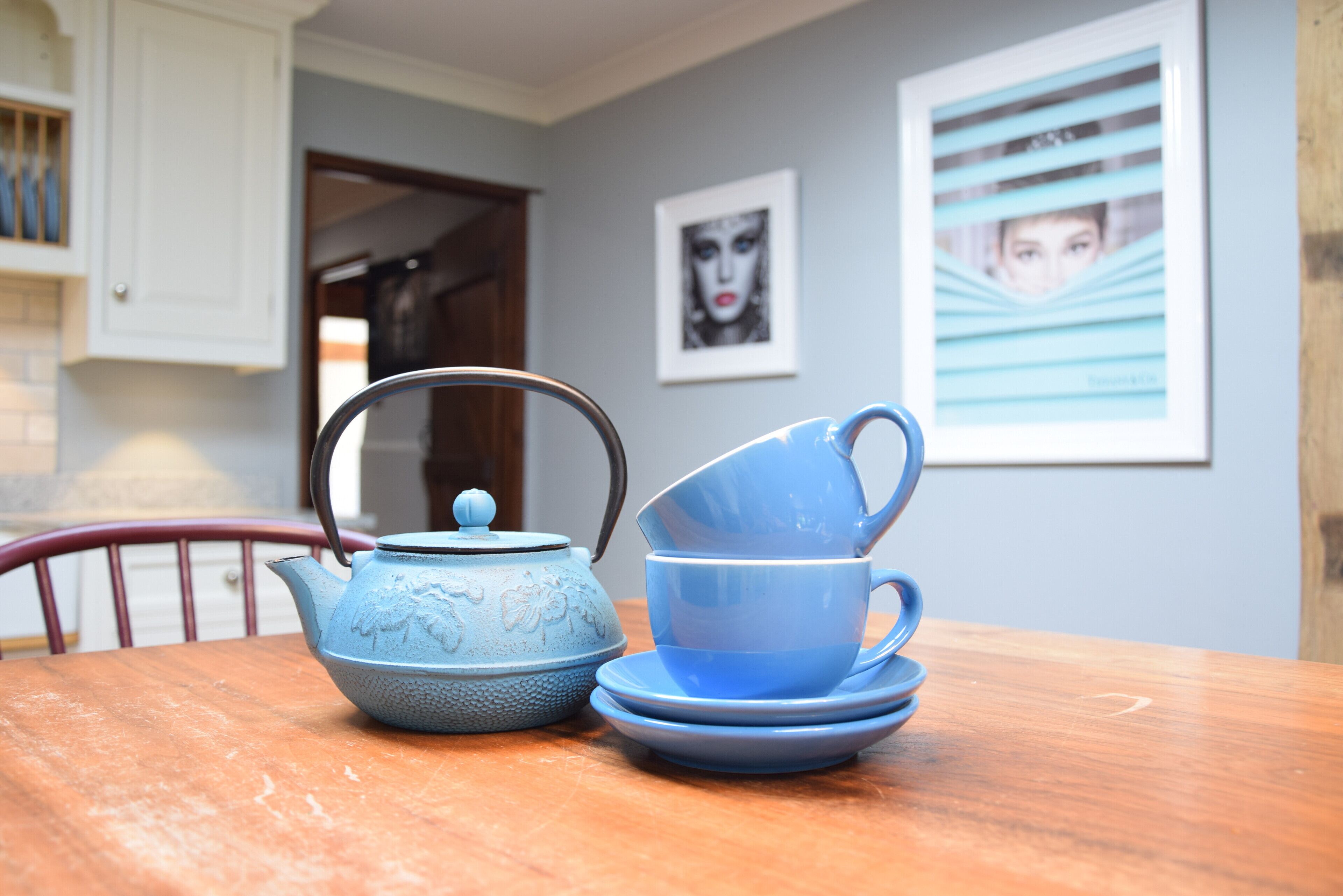 A blue teapot and cup set on a wooden table in a room with a door and framed pictures on the wall.