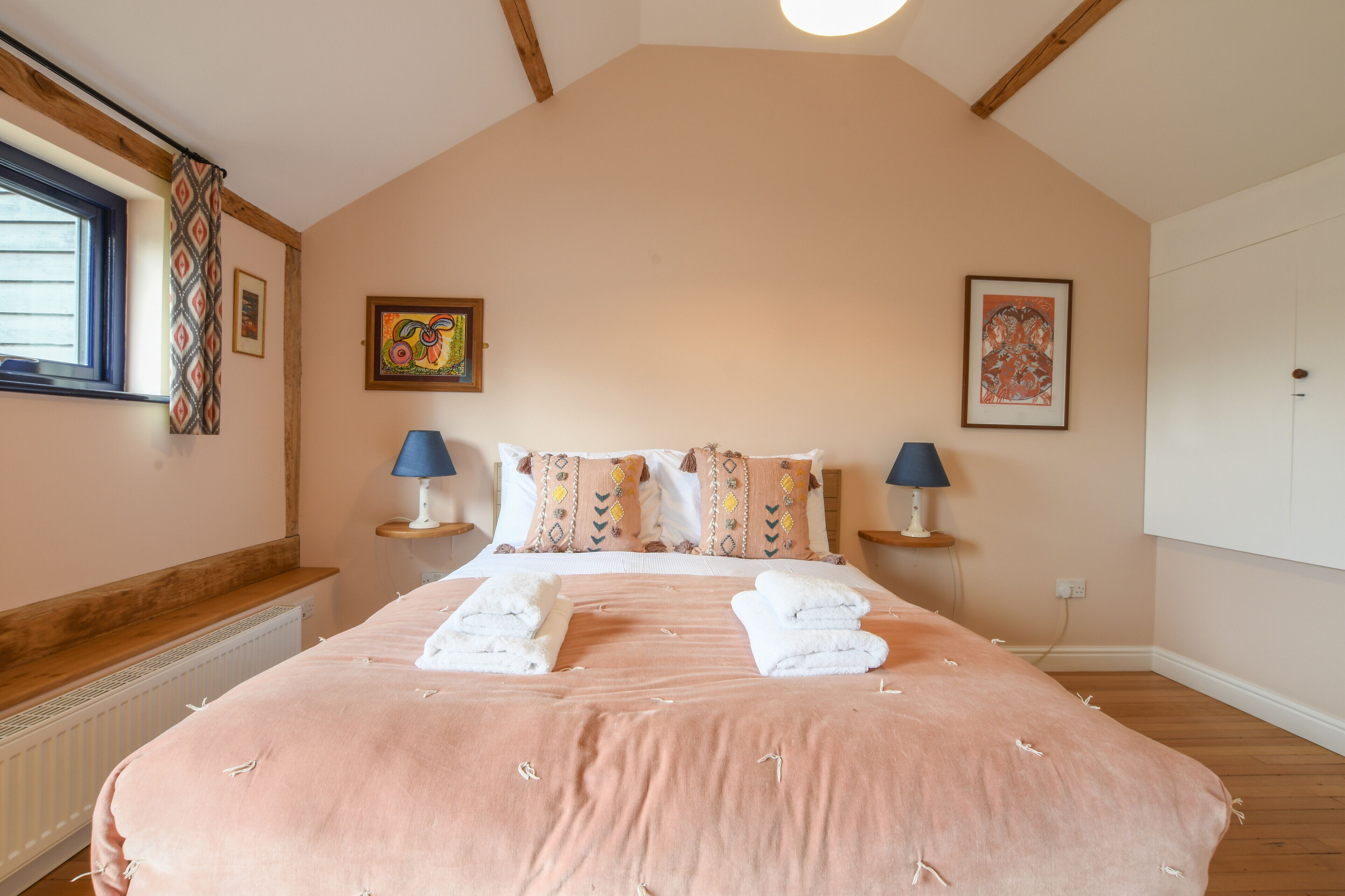 Une chambre avec un grand lit, deux tables de chevet avec des lampes, une fenêtre avec des rideaux à motifs et deux œuvres d'art encadrées accrochées aux murs.