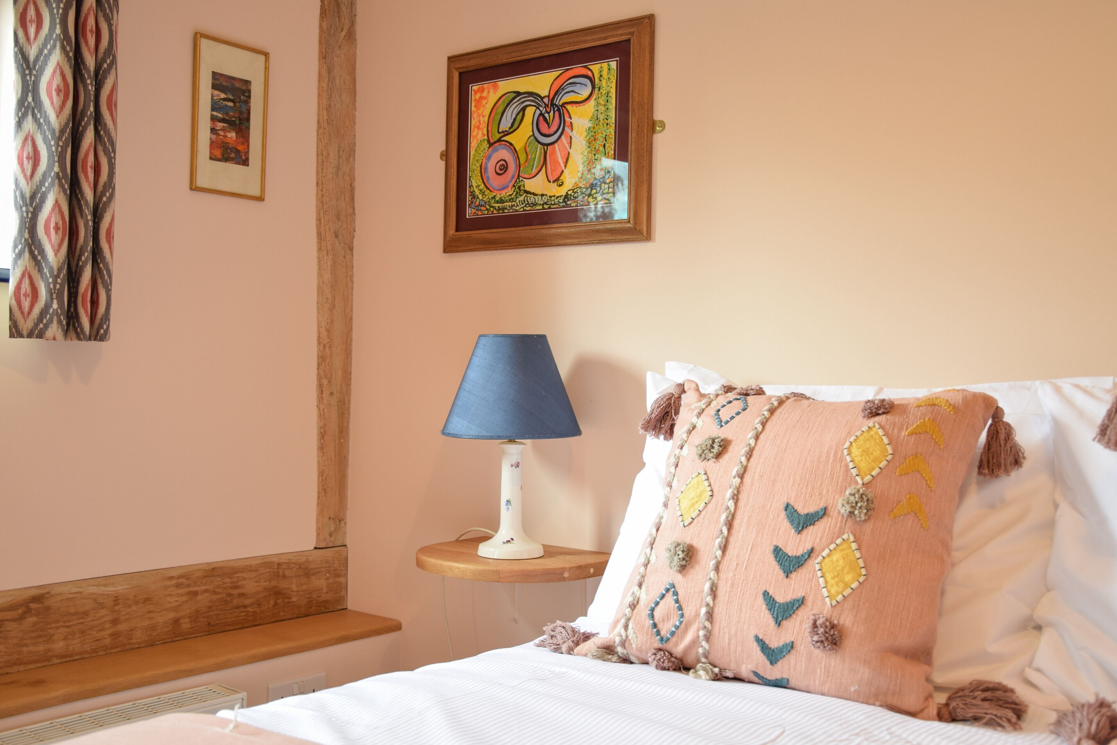 A neatly made bed with a blue lamp and a colorful pillow, a framed painting on the wall, and a patterned curtain.