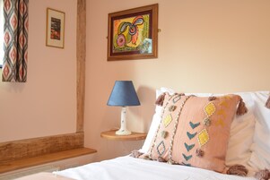 A neatly made bed with a blue lamp and a colorful pillow, a framed painting on the wall, and a patterned curtain.