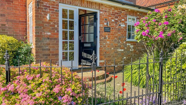Une petite maison en briques avec une porte noire, une enseigne indiquant « CHAPEL COTTAGE », et un jardin agrémenté de diverses fleurs et d’une clôture en métal.