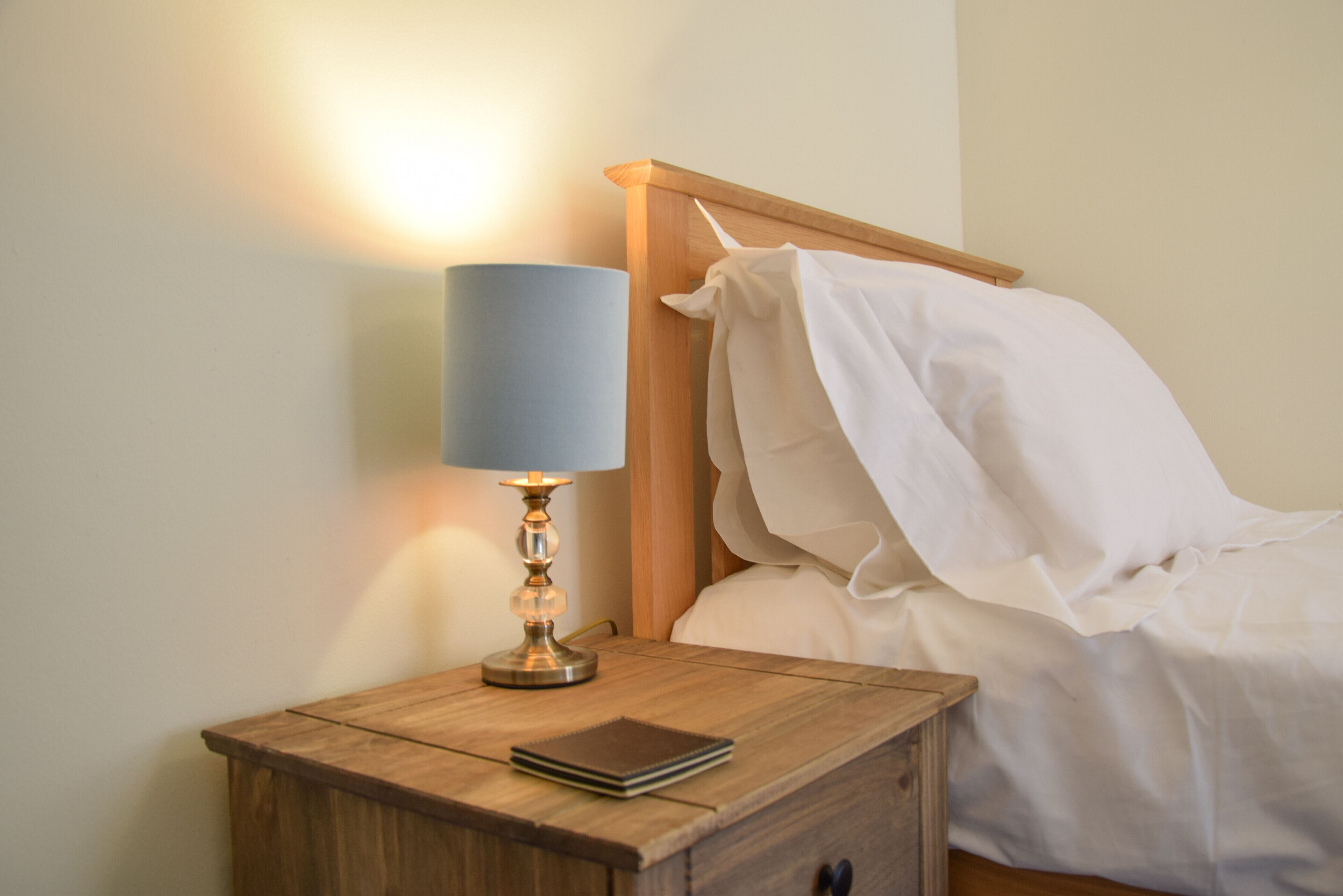 A wooden bedside table with a lamp, a notebook, and a bed with white linens.