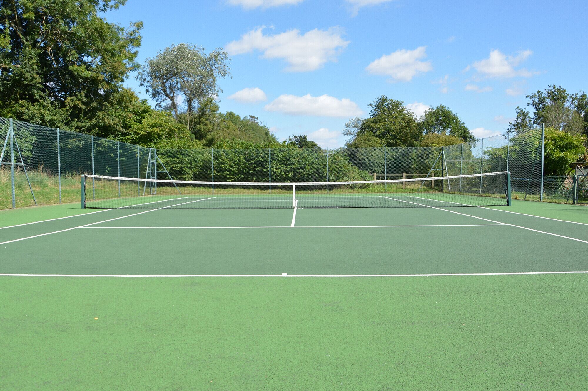 An outdoor tennis court with a net, surrounded by a green fence and trees.
