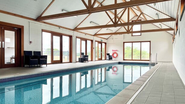 An indoor swimming pool with a tiled floor and clear blue water. There are wooden beams on the ceiling and large windows on the walls. - The Gallery, Blythburgh (Halesworth)