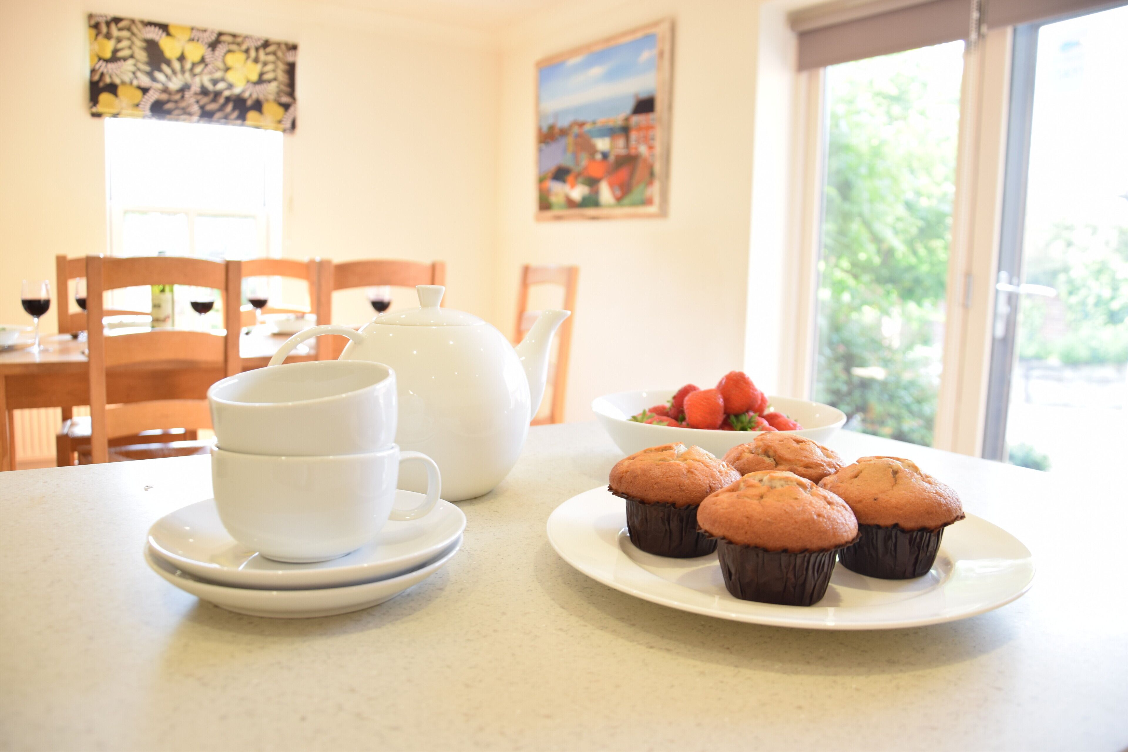 A breakfast table with white cups, a teapot, a plate of muffins, and a bowl of strawberries.