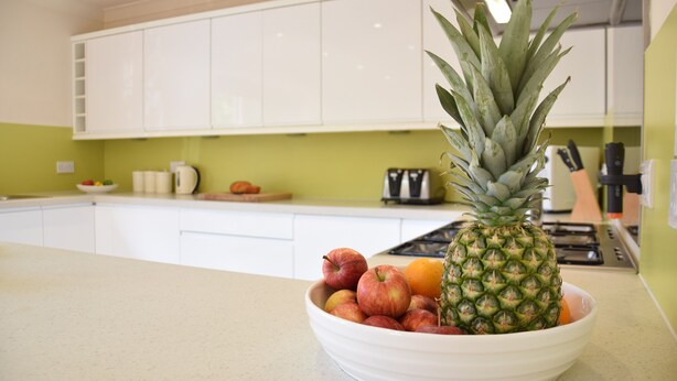 Une cuisine avec des meubles blancs, un saladier rempli de fruits et un ananas posé sur le comptoir.