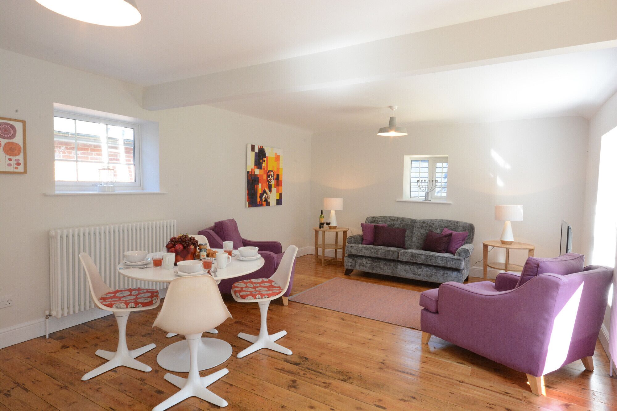 A modern living room with a round dining table, two purple armchairs, a grey sofa, and a white radiator.