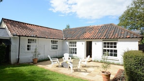 Ein weißes Cottage mit Ziegeldach, einer kleinen Terrasse mit weißen Stühlen und einem Garten.