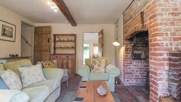 A cozy living room with a brick fireplace, a wooden cabinet, and a green sofa. - Shoemakers Cottage, Friston (Saxmundham)