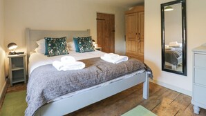 A neatly made bed with pillows, a bedside lamp, a wooden wardrobe, and a mirror on the wall. - Shoemakers Cottage, Friston (Saxmundham)