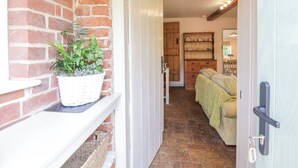A narrow hallway with a brick wall on one side and a wooden door on the other. There is a white shelf with a basket of green plants. - Shoemakers Cottage, Friston (Saxmundham)