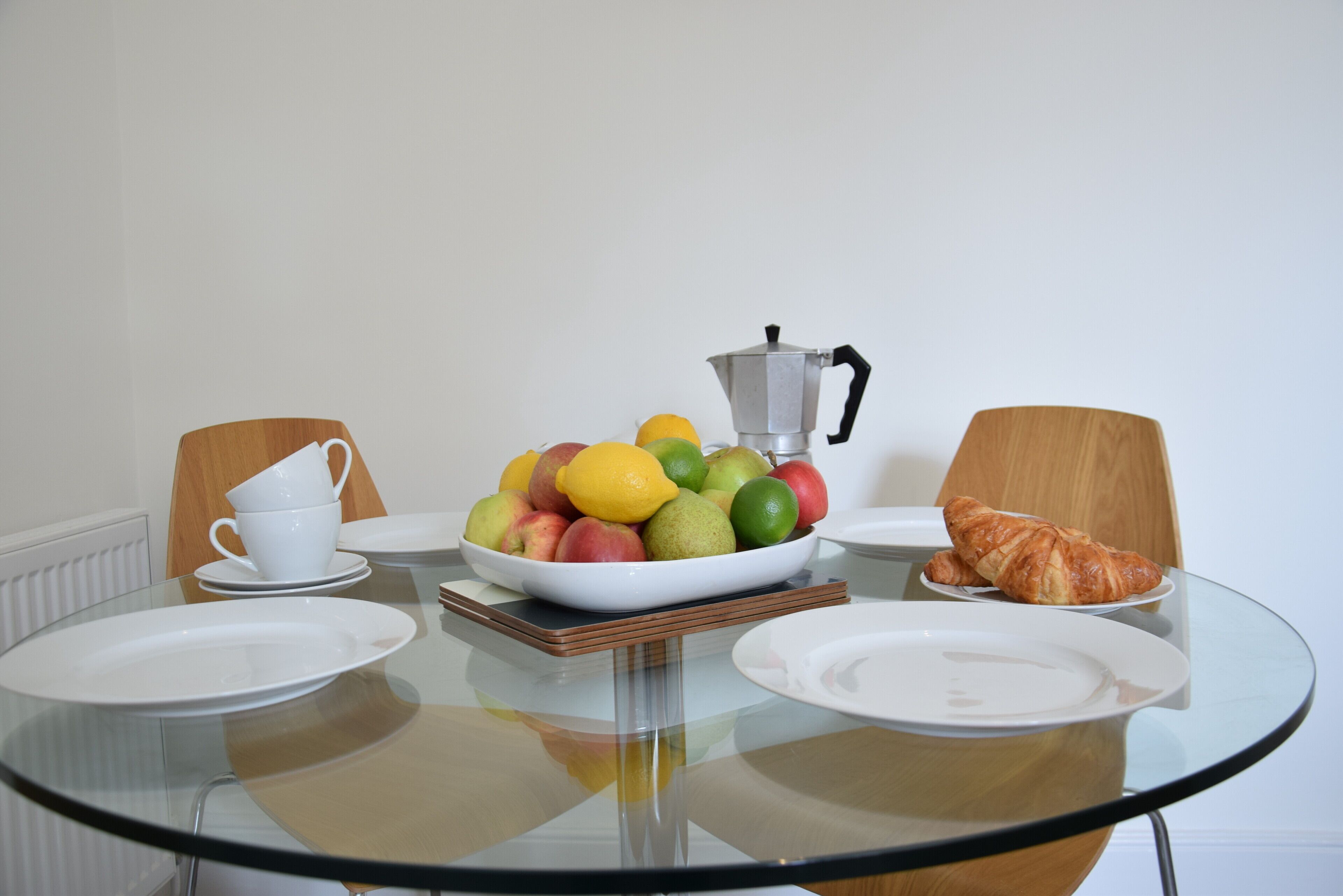Ein Frühstückstisch mit Obstschale, Kaffeekanne, Croissant und weißen Tellern.