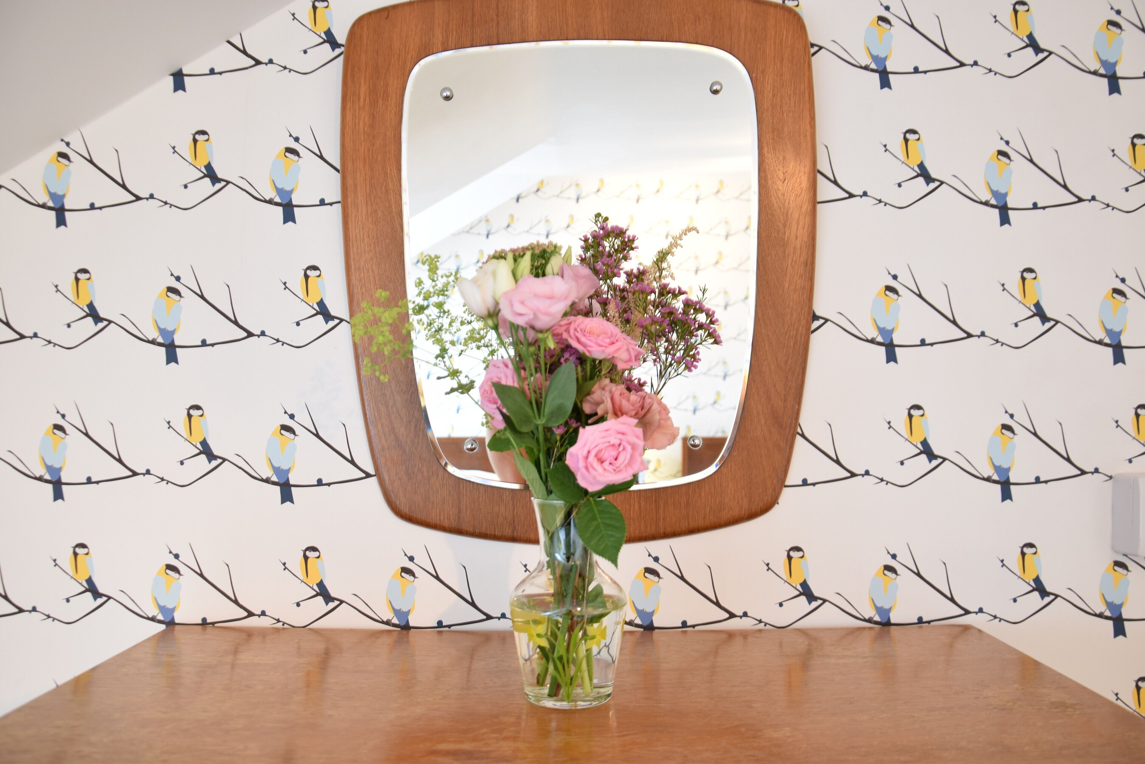 A wooden table with a clear glass vase containing pink flowers, set against a wall with a bird pattern wallpaper.