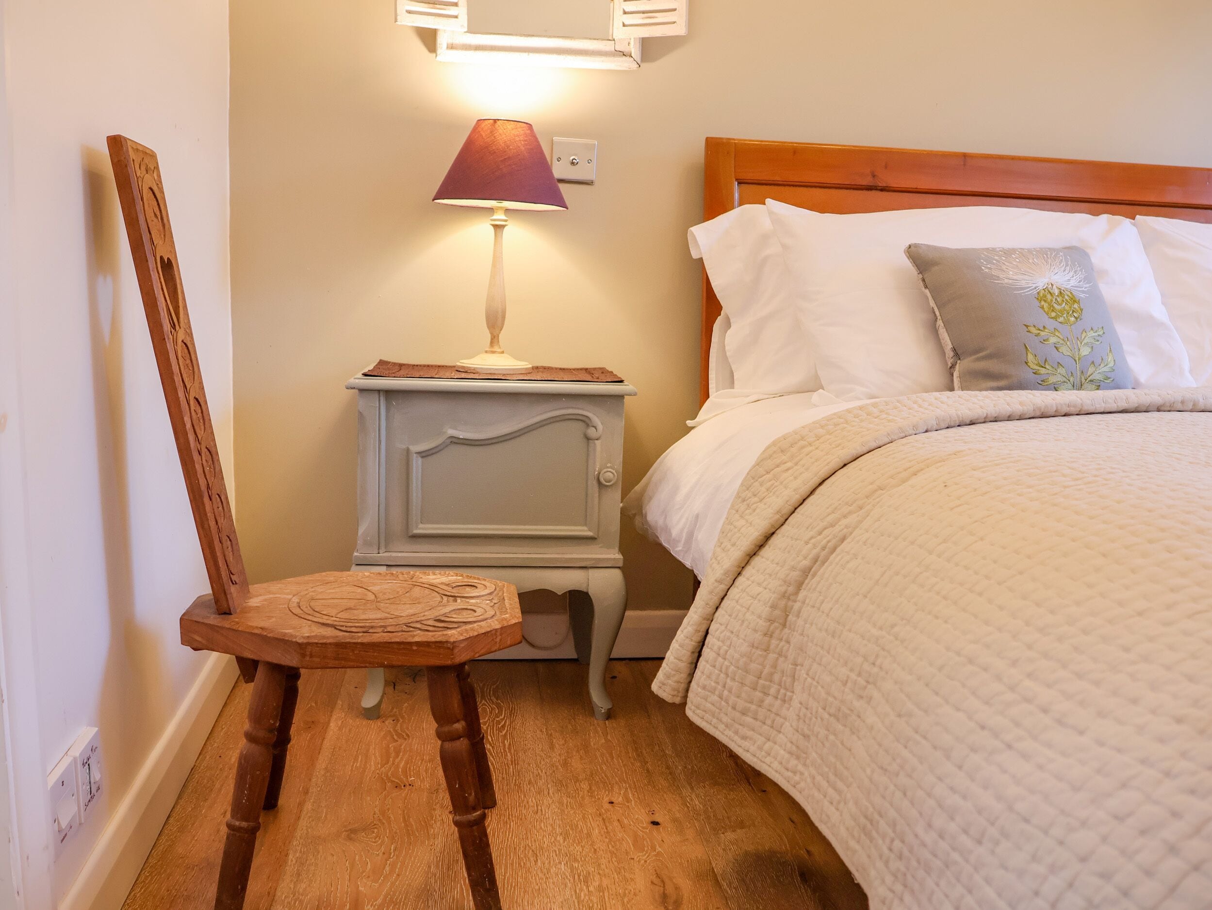 A bedroom with a wooden chair, a bedside table with a lamp, a bed with white bedding, and a nightstand.