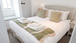En snyggt bäddad säng med vita sängkläder och en grön och beige pläd, två kuddar med ett geometriskt mönster och en sänglampa på ett litet bord bredvid sängen.