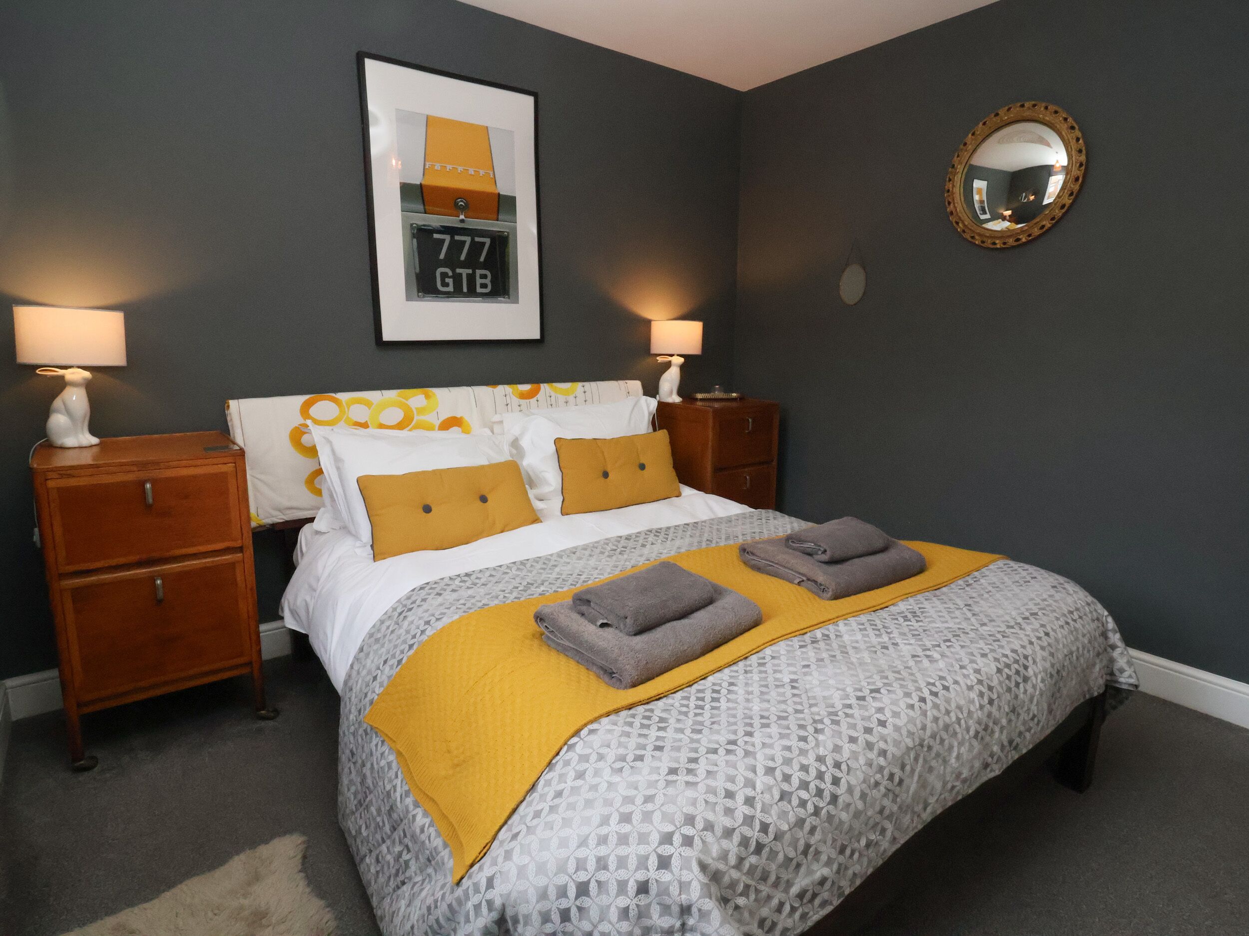 Een opgemaakt bed met een beddekleed met patroon, twee kussens en een gevouwen handdoek. Er staan twee nachtkastjes met lampen, een ingelijste foto aan de muur en een spiegel.
