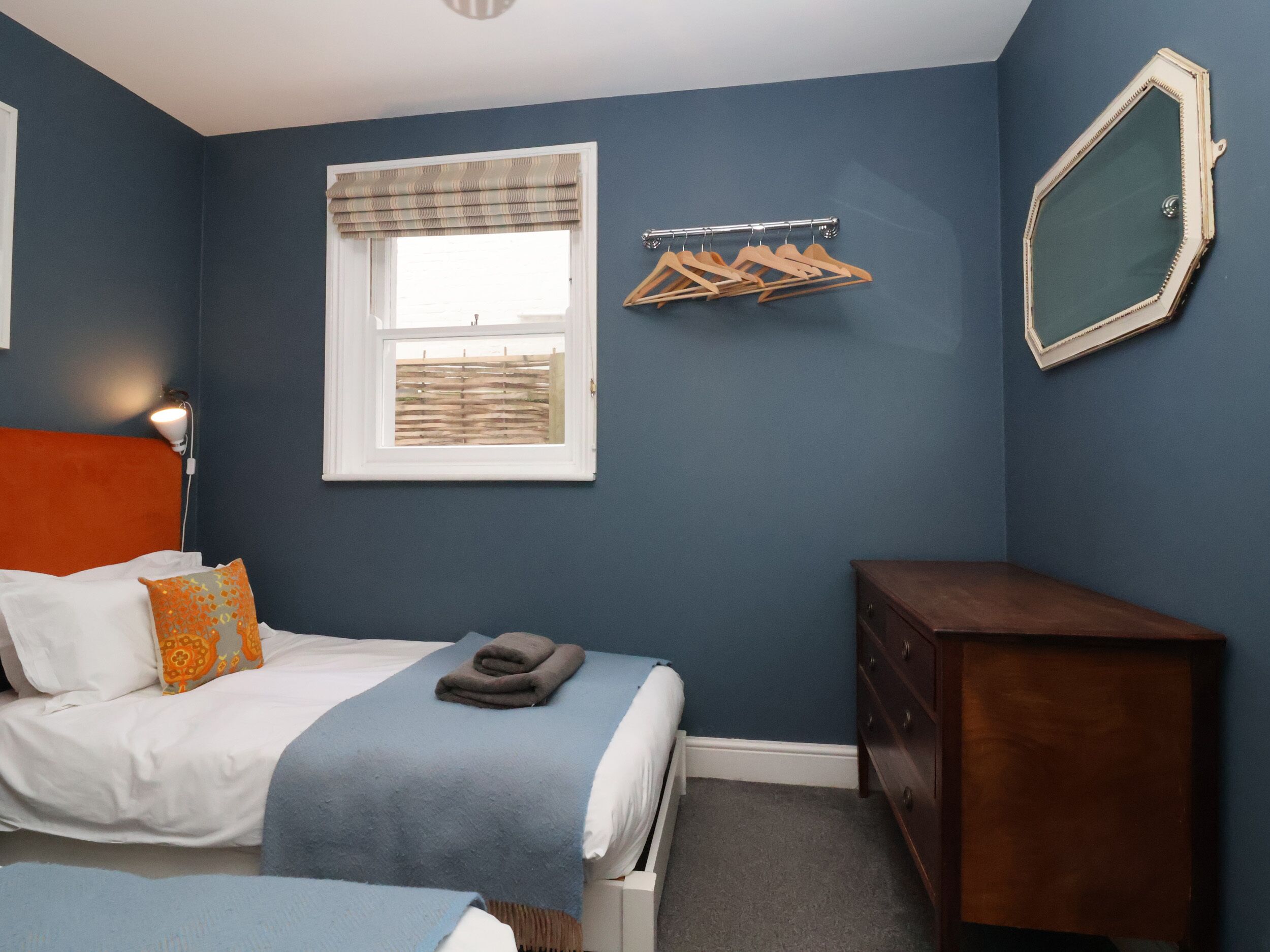 A bedroom with a bed, a dresser, a window with blinds, a wall-mounted mirror, and a clothes rack.