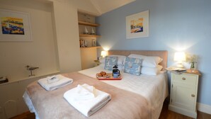 A neatly made bed with pillows, a bedside table with a lamp, and a shelf with books and decorative items. - Garden Cottage, Southwold (Reydon)