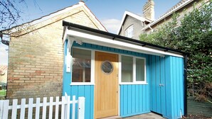 A small, single-story building with a blue exterior, a wooden door, and a white picket fence in front. - Garden Cottage, Southwold (Reydon)