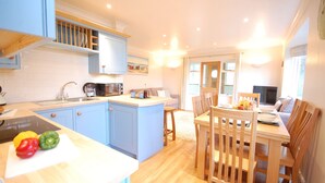 A modern kitchen with blue cabinets, a coffee machine, and a dining area with wooden chairs and a table. - Garden Cottage, Southwold (Reydon)